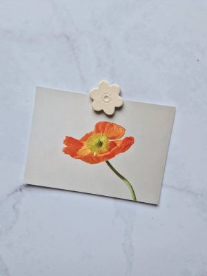 Magnet en grès émaillé écru en forme de fleur posé sur une carte postale