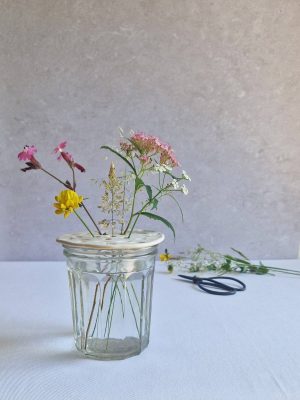 Pique-fleurs posé sur un verre rempli d'eau et contenant des fleurs sauvages, posé à côté de ciseaux de jardinier